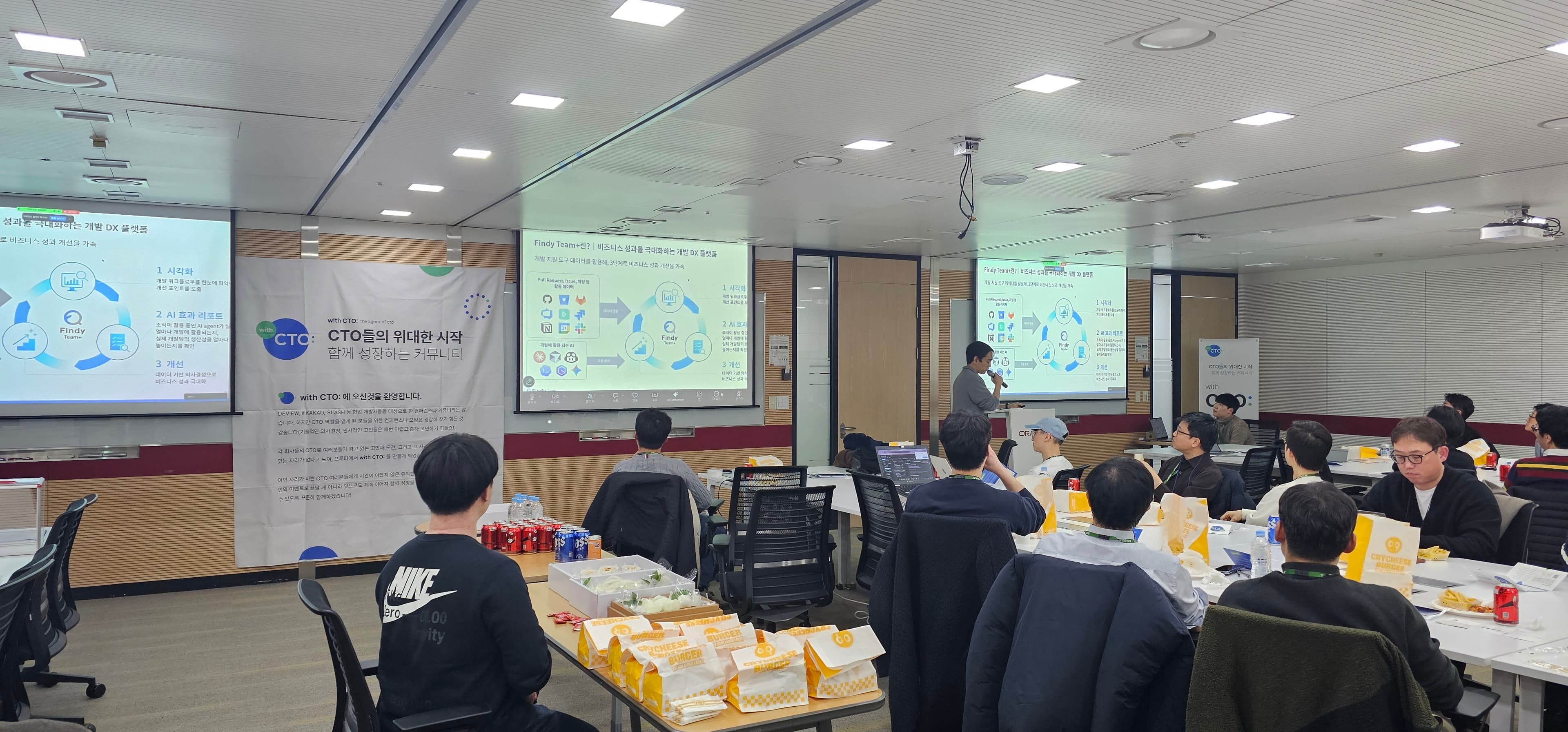 Findy Team+ 개발 DX 플랫폼 슬라이드를 띄우고 발표자가 말하는 회의실, 앞쪽에 크라이치즈버거 봉투가 쌓여 있음