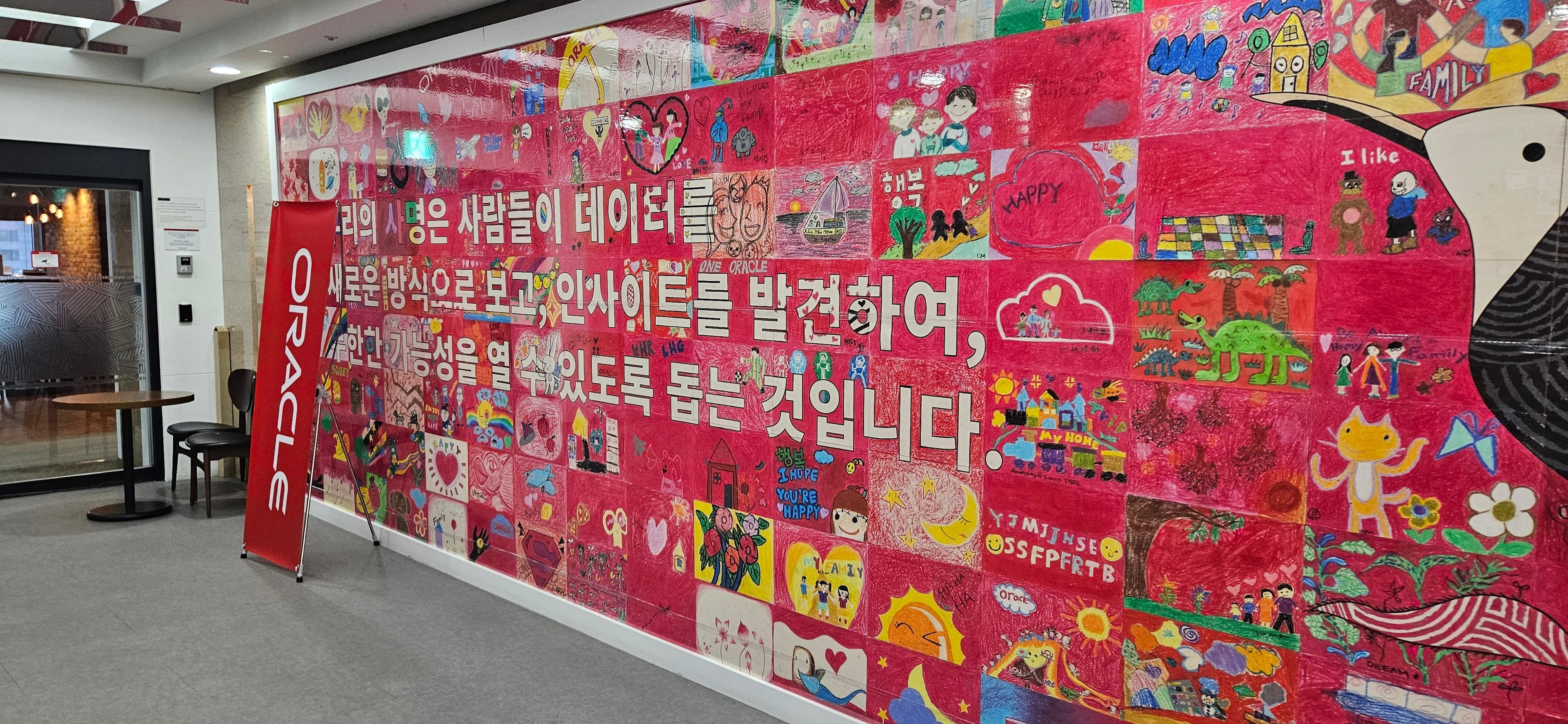 아이들 그림으로 채운 붉은 벽에 오라클의 사명 문구가 적혀 있고 앞에 ORACLE 배너가 선 로비