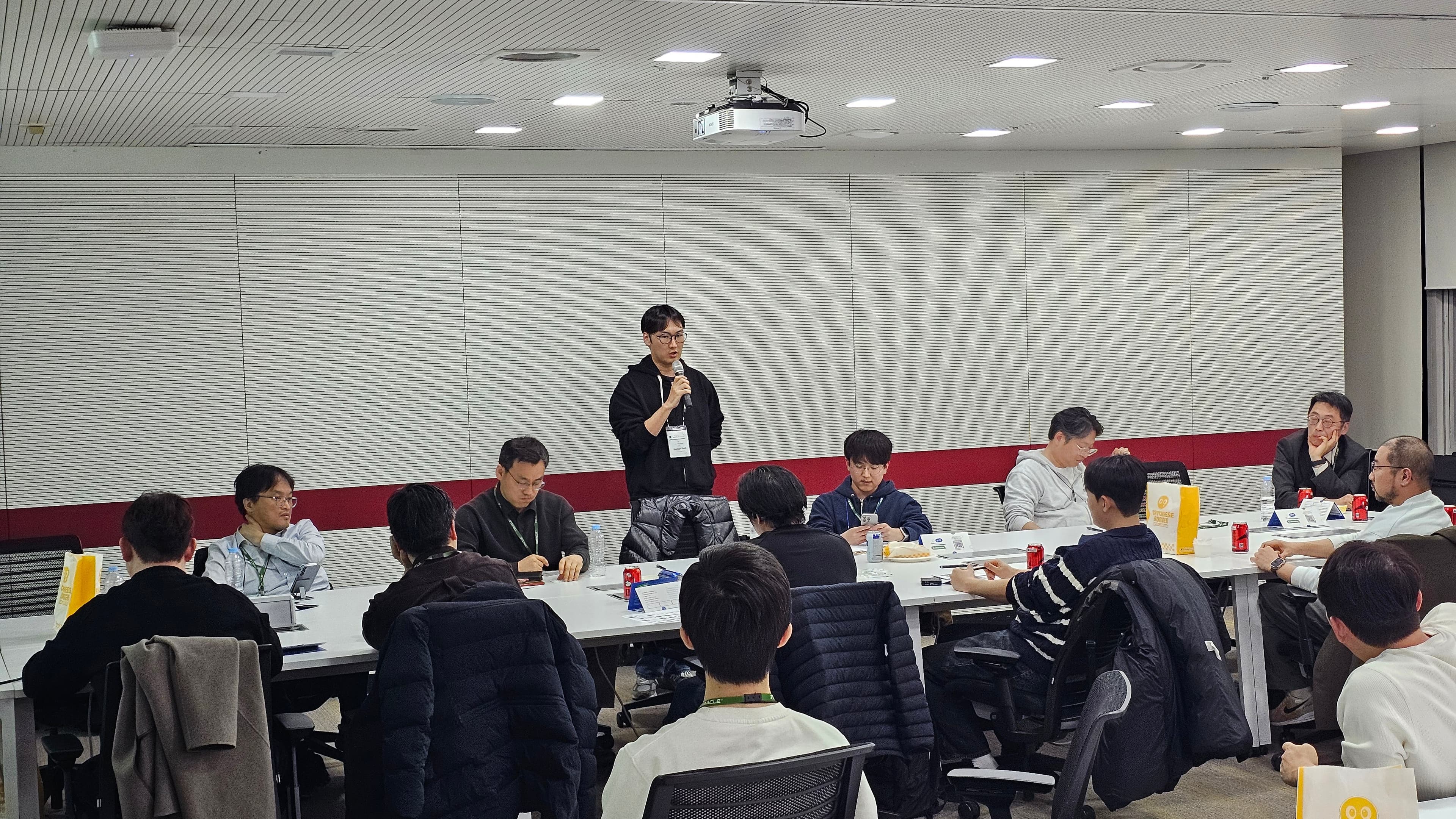 디귿 자로 놓인 테이블에 앉은 참석자들 앞에서 한 사람이 서서 마이크로 이야기하는 장면