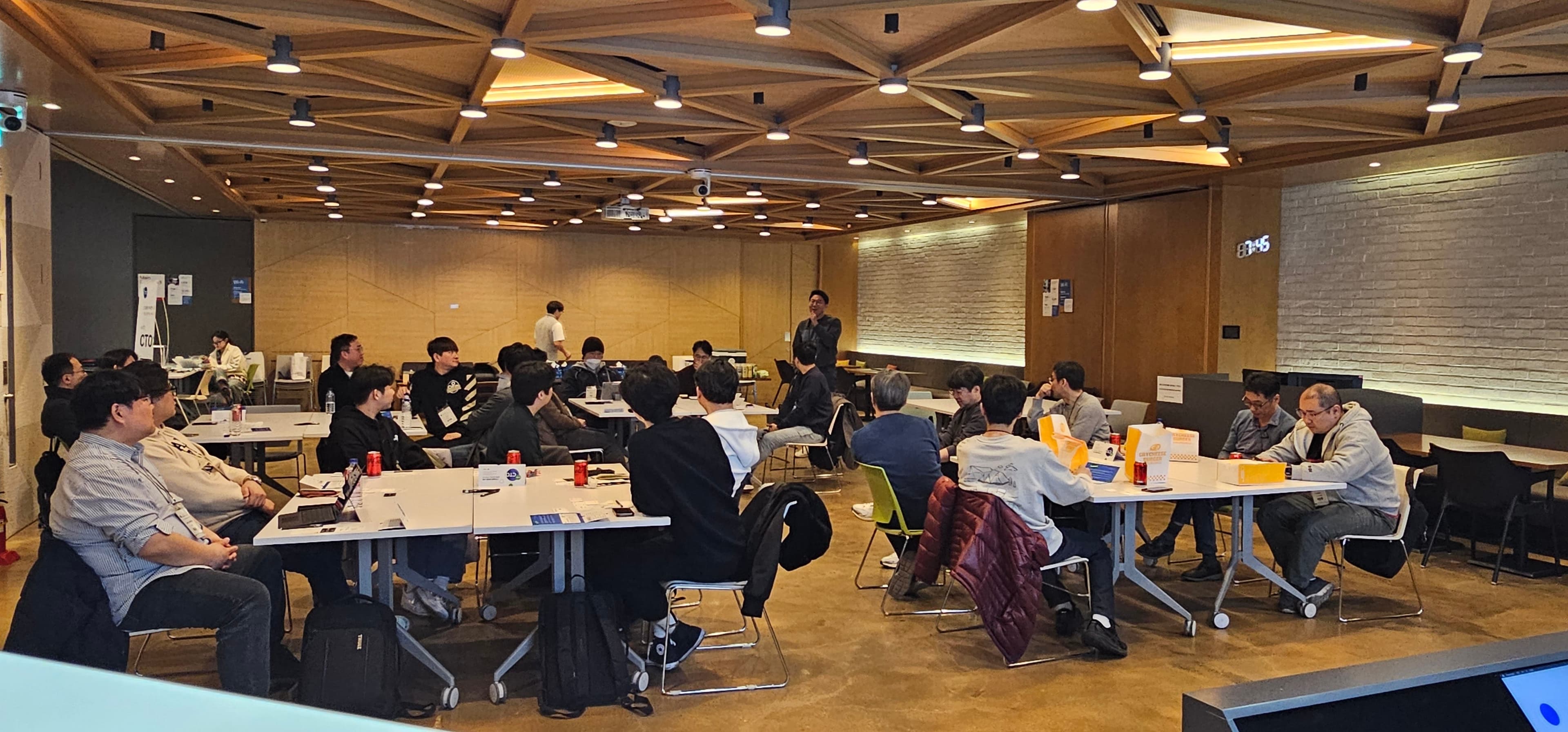 크라이치즈버거 상자가 놓인 테이블에 앉은 참가자들과 일어서서 말하는 사람