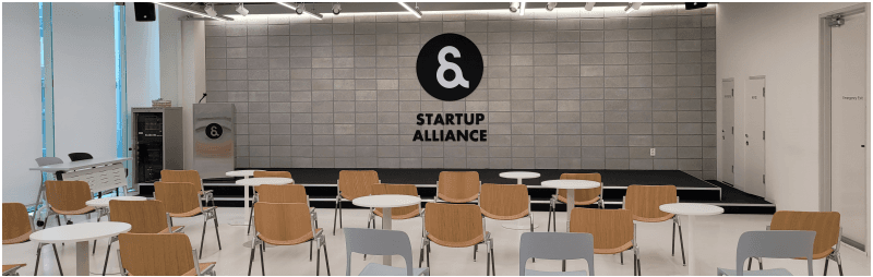 STARTUP ALLIANCE 로고가 걸린 무대와 원형 테이블, 의자가 놓인 빈 행사장