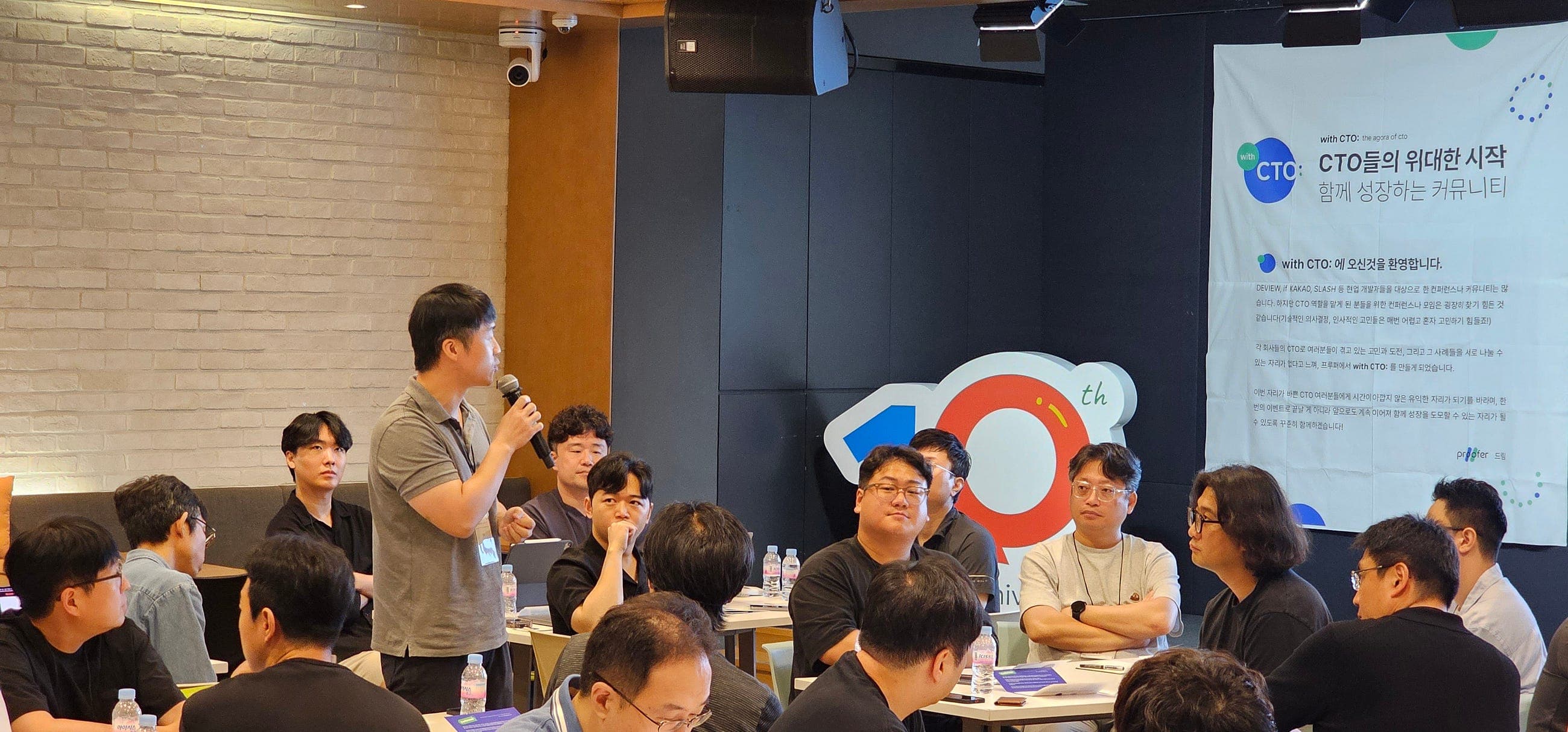한 참여자가 일어서서 마이크로 말하고 테이블의 참여자들이 듣는 토크쇼 장면