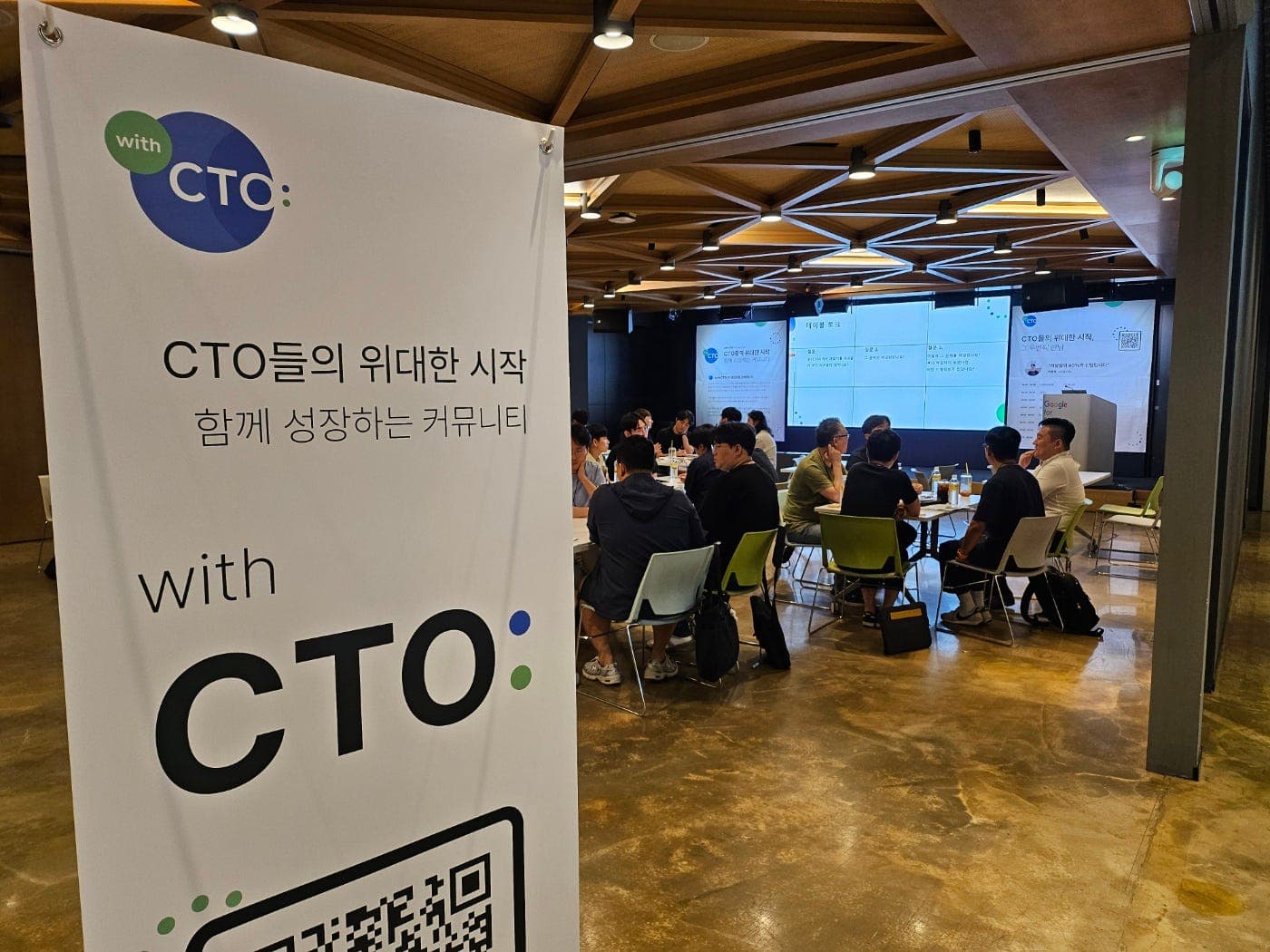 CTO들의 위대한 시작 문구가 적힌 with CTO: 배너 뒤로 참여자들이 테이블에 둘러앉아 대화하는 행사장