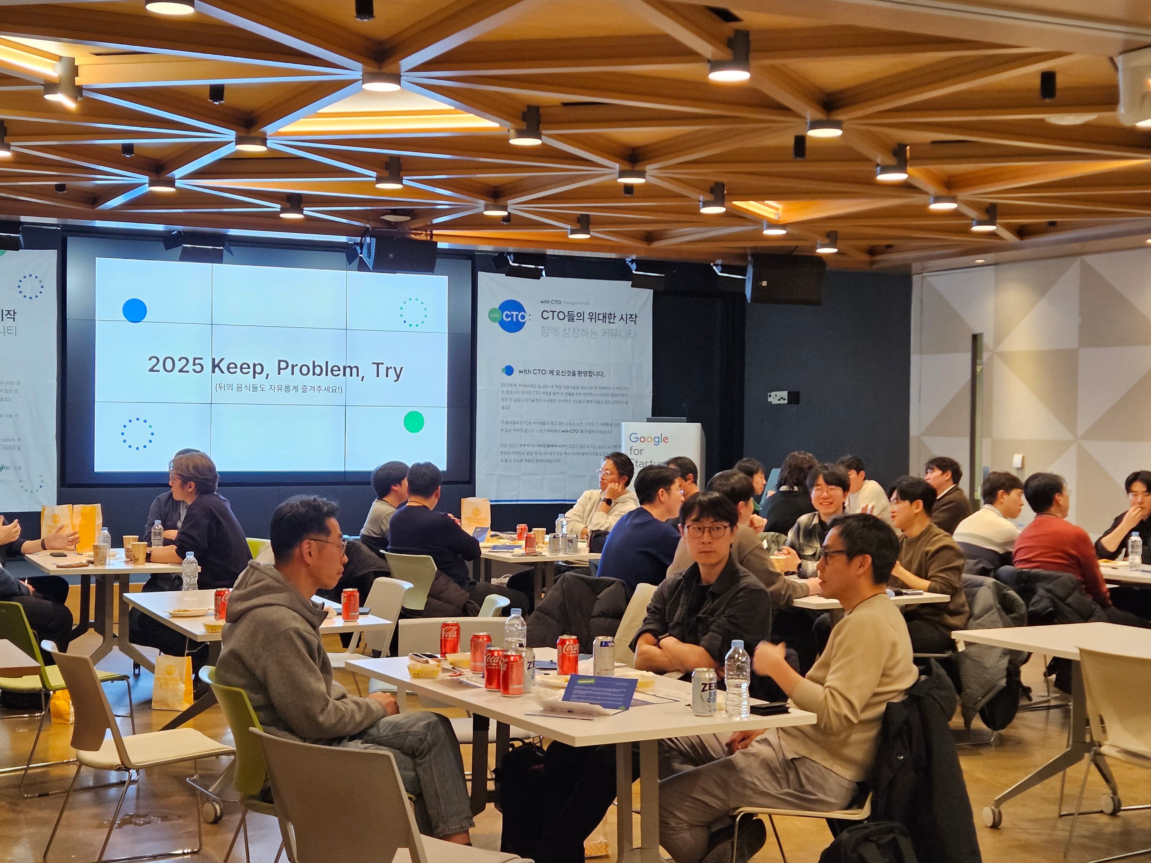'2025 Keep, Problem, Try' 화면 앞 테이블에 콜라와 버거 봉투를 두고 대화하는 참가자들