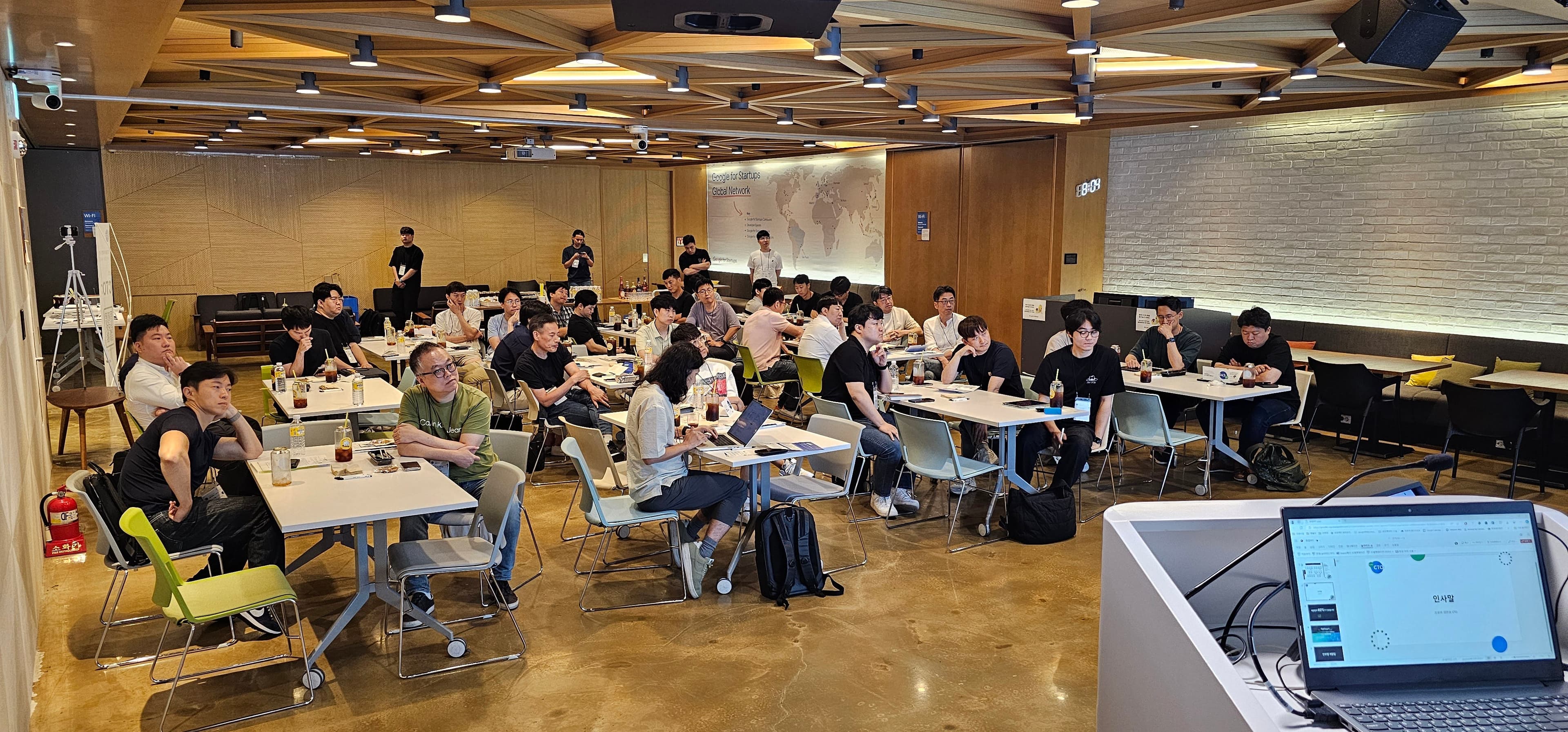 연단 노트북에 '인사말' 화면이 열려 있고 테이블에 앉아 앞을 바라보는 참가자들